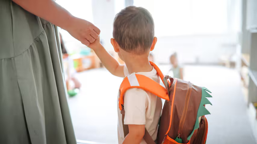 A young child wearing a backpack holds an adult's hand, preparing to enter a bright, indoor space, possibly a classroom or school.